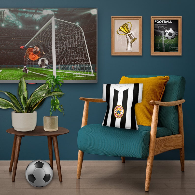 May include: A teal armchair with wooden arms and legs, a black and white striped throw pillow with a star logo, and a yellow throw pillow. The chair is in front of a blue wall with two framed pictures of a trophy and a football game.