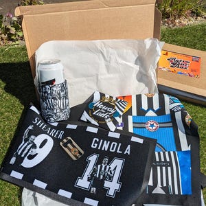 May include: An open cardboard box containing a Newcastle United football-themed gift set. Includes a black and white can cooler, a black and white towel with player names and numbers, and a jersey-style scarf. A Retro Star Box sticker is visible.