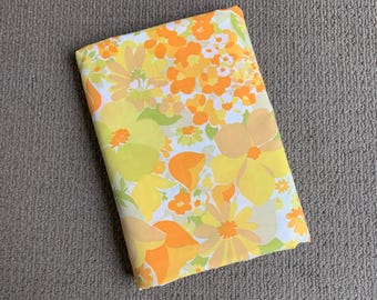 May include: A folded piece of fabric featuring a vibrant floral pattern with various shades of yellow, orange, and light green flowers on a white background. The textile displays a dense arrangement of blossoms and leaves in a repeating design.