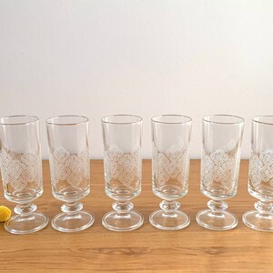 May include: Six clear glass drinking glasses with a white floral design. The glasses have a short stem and a wide base.