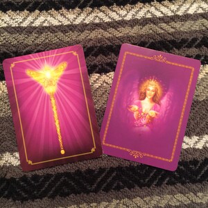 Puede incluir: Dos cartas de tarot moradas con bordes dorados. La carta de la izquierda presenta una varita dorada con una mariposa en la parte superior. La carta de la derecha presenta un &aacute;ngel con alas y un halo dorado.
