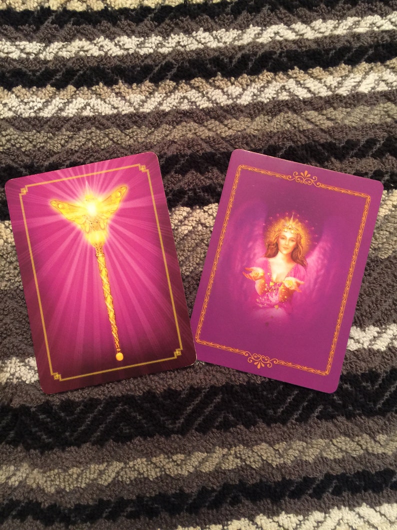Puede incluir: Dos cartas de tarot moradas con bordes dorados. La carta de la izquierda presenta una varita dorada con una mariposa en la parte superior. La carta de la derecha presenta un &aacute;ngel con alas y un halo dorado.