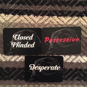 Puede incluir: Tres cartas negras con texto blanco y un dise&ntilde;o de rosa en blanco y negro. El texto en las cartas dice "Closed Minded", "Possessive" y "Desperate".
