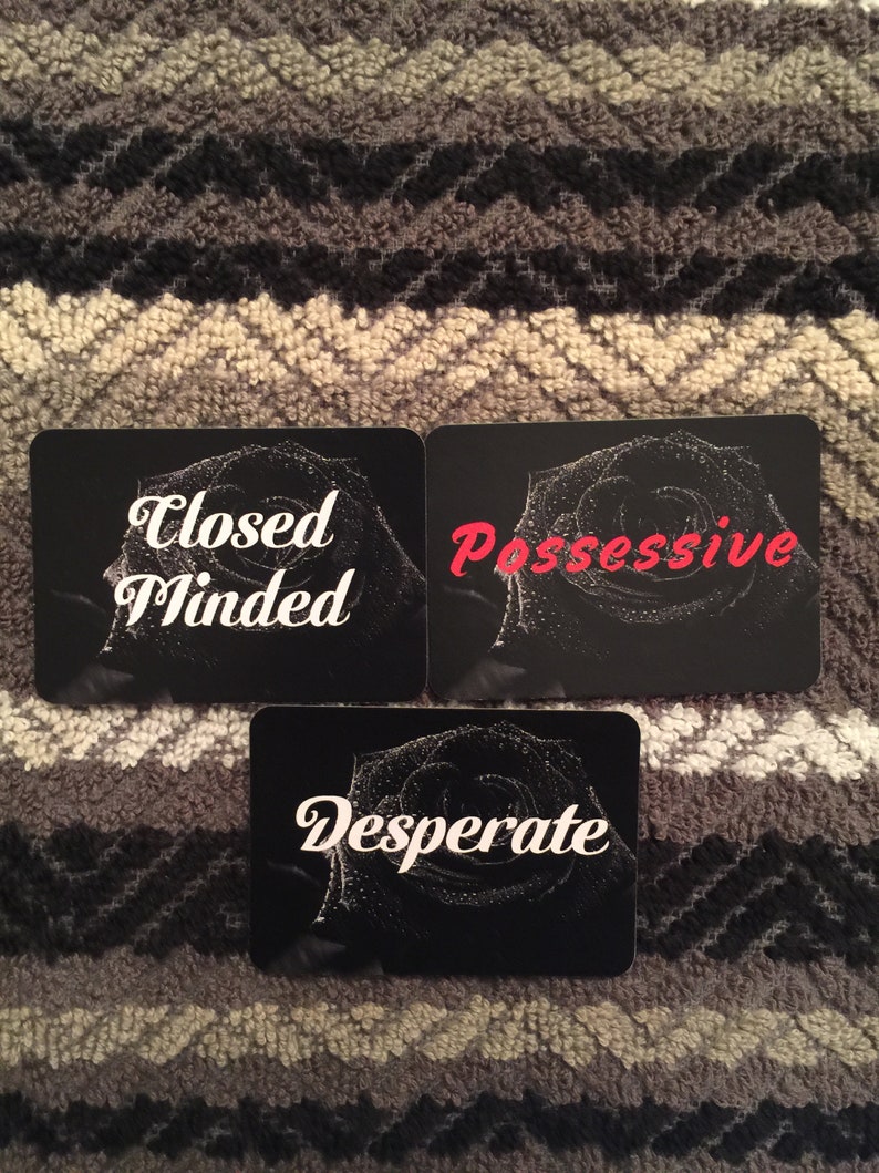 Puede incluir: Tres cartas negras con texto blanco y un dise&ntilde;o de rosa en blanco y negro. El texto en las cartas dice "Closed Minded", "Possessive" y "Desperate".