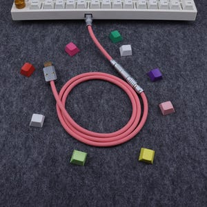 May include: A pink coiled USB cable with silver connectors is connected to a white keyboard. Scattered around the cable are colorful keyboard keycaps in various colors, including pink, green, and purple. The background is a dark gray fabric.