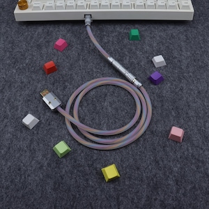 May include: A coiled, multi-colored USB cable with a silver connector and a white keyboard in the background. Scattered around the cable are colorful keycaps in various shades, including pink, green, and purple. The cable has a pastel rainbow design.