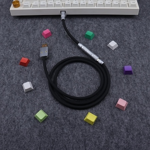 Könnte beinhalten: Eine weiße mechanische Tastatur mit einem gewickelten schwarzen Kabel und einem silbernen Stecker. Bunte Tastenkappen in Pink, Grün, Rot, Weiß, Gelb und Lila sind verstreut. Die Tastatur hat einen goldfarbenen Knopf.