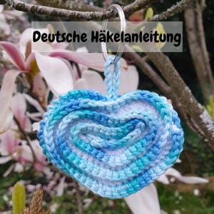 May include: A blue and white crocheted heart-shaped keychain with a silver key ring. The text "Deutsche Häkelanleitung" is visible above the keychain.