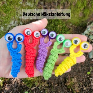 May include: A hand holds six colorful, crocheted worm-like figures with large googly eyes. The worms are blue, red, purple, green, and yellow. The text "Deutsche Häkelanleitung" is at the top.