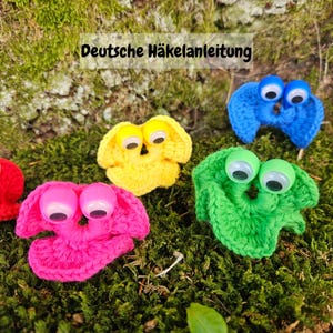 May include: A collection of colorful, crocheted creatures with large, googly eyes. The creatures are in shades of pink, yellow, green, and blue, and are set against a backdrop of green moss. The text "Deutsche Häkelanleitung" is visible.
