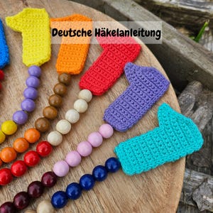 Op de afbeelding: Een regenboogkleurige gehaakte vingerpopenslinger met houten kralen. De slinger ligt op een houten oppervlak. De tekst "Deutsche Häkelanleitung" is zichtbaar in de afbeelding.