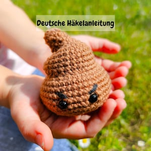 Könnte beinhalten: Ein gehäkeltes braunes Kack-Emoji mit schwarzen Augen und Augenbrauen. Der Text "Deutsche Häkelanleitung" befindet sich über dem Emoji.