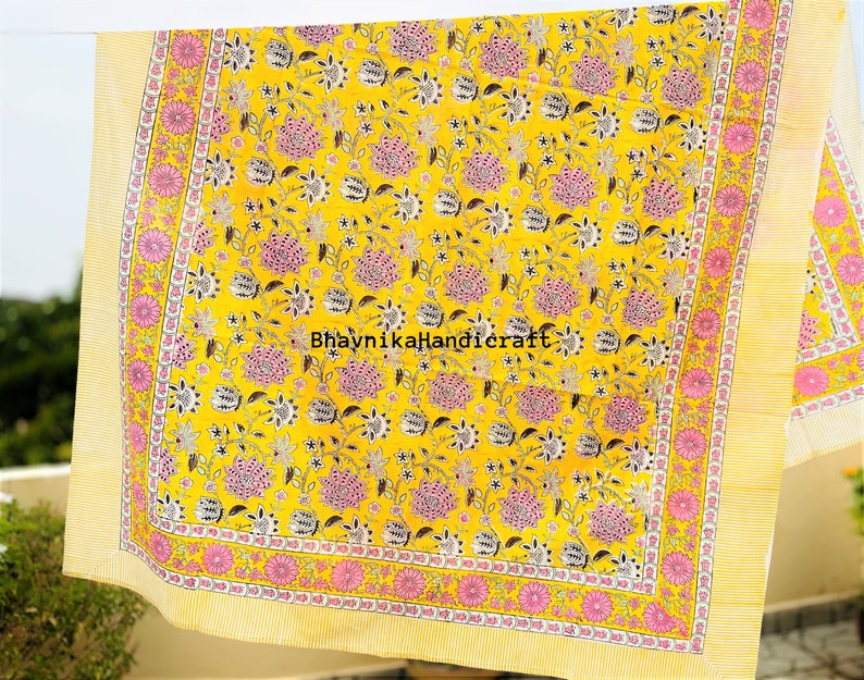 May include: Yellow cotton fabric with a pink and black floral print. The fabric has a yellow border with a pink and white floral pattern. The text "Bhavnika Handicraft" is printed in the center of the fabric.
