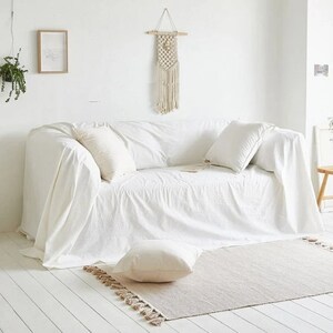 Peut inclure: Un canap&eacute; blanc avec une couverture blanche et deux coussins blancs. Le canap&eacute; est dans une pi&egrave;ce blanche avec un tapis blanc et une suspension murale en macram&eacute;.