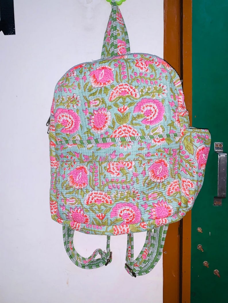 Puede incluir: Una mochila de color azul claro y rosa con estampado floral y correas verdes. La mochila tiene un dise&ntilde;o acolchado y una cremallera.