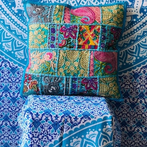 Op de afbeelding: Een turquoise blauwe kussen met een patchwork ontwerp met kleurrijke geborduurde bloemenpatronen en pailletten. Het kussen ligt op een blauw-wit gepatroneerd oppervlak.