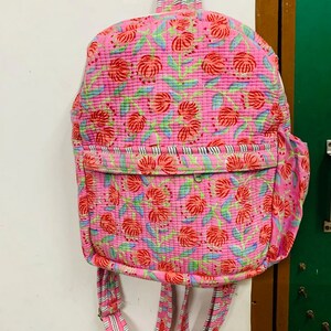 Puede incluir: Una mochila rosa con estampado floral y correas blancas y negras a rayas. La mochila tiene un peque&ntilde;o bolsillo en la parte delantera.
