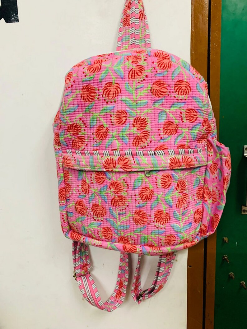 Puede incluir: Una mochila rosa con estampado floral y correas blancas y negras a rayas. La mochila tiene un peque&ntilde;o bolsillo en la parte delantera.