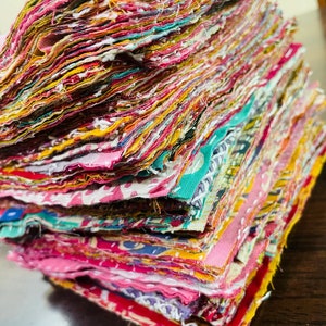 Pode incluir: Uma pilha de quadrados de tecido coloridos, cada um com um padrão diferente, estão empilhados cuidadosamente sobre uma superfície de madeira. Os quadrados de tecido são uma mistura de rosa, amarelo, verde e azul.