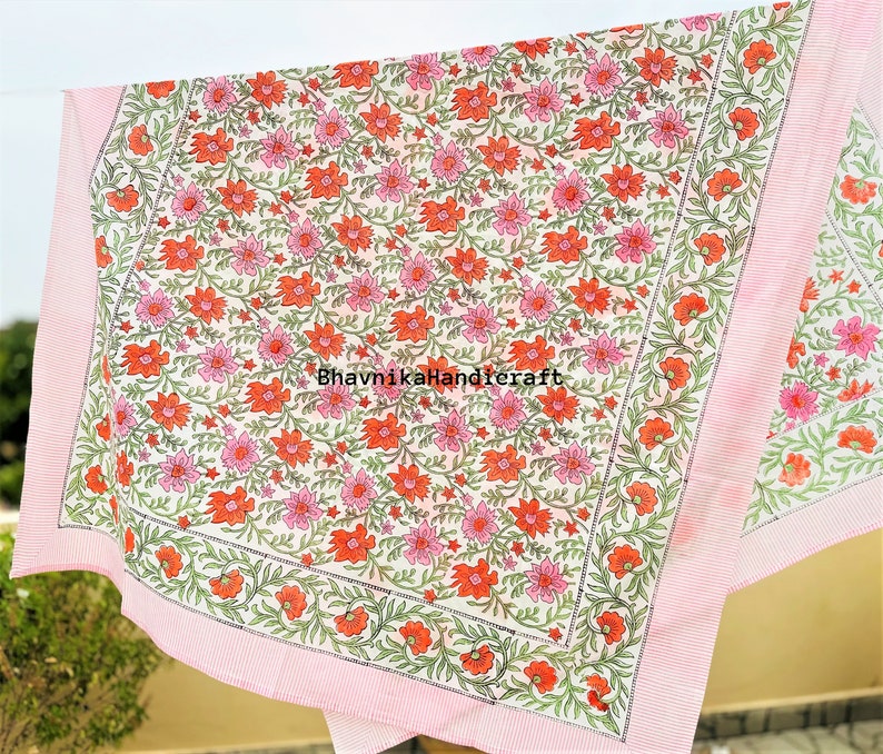 May include: A white cotton tablecloth with a pink border and a floral print featuring pink and orange flowers. The tablecloth is printed with the text "BhavnikaHandicraft".