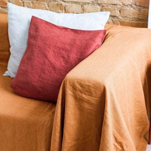 Peut inclure: Un jet&eacute; en lin brun drap&eacute; sur un canap&eacute; avec un coussin rouge et un coussin blanc. Le canap&eacute; est devant un mur en briques.