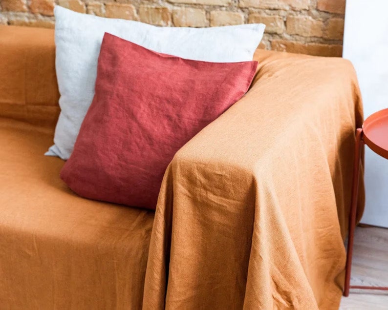 Peut inclure: Un jet&eacute; en lin brun drap&eacute; sur un canap&eacute; avec un coussin rouge et un coussin blanc. Le canap&eacute; est devant un mur en briques.