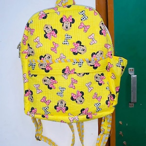 Puede incluir: Una mochila acolchada amarilla con estampado de Minnie Mouse. La mochila tiene cierre de cremallera y correas ajustables.