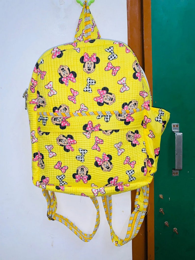 Puede incluir: Una mochila acolchada amarilla con estampado de Minnie Mouse. La mochila tiene cierre de cremallera y correas ajustables.