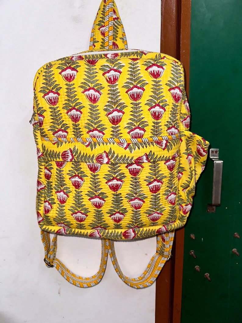 Puede incluir: Una mochila acolchada amarilla con un patr&oacute;n floral. La mochila tiene flores y hojas rojas y verdes sobre un fondo amarillo. Las correas son de rayas amarillas y blancas.