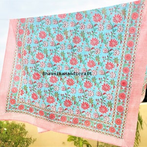 May include: A light blue cotton fabric with a pink floral print. The fabric has a pink border with a white and pink pattern. The fabric is folded and hanging on a clothesline. The text "BhavnikaHandicraft" is printed on the fabric.