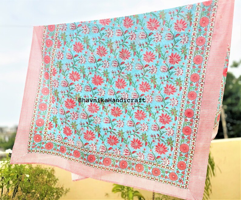 May include: A light blue cotton fabric with a pink floral print. The fabric has a pink border with a white and pink pattern. The fabric is folded and hanging on a clothesline. The text "BhavnikaHandicraft" is printed on the fabric.
