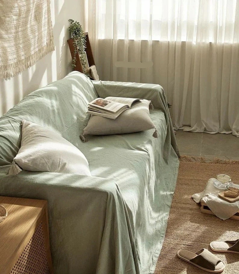 Peut inclure: Une couverture de canap&eacute; en tissu l&eacute;ger vert clair avec des glands sur les bords. La couverture est drap&eacute;e sur un canap&eacute; vert clair avec des coussins blancs. Le canap&eacute; est dans un salon avec une table d'appoint en bois et un tapis au sol.