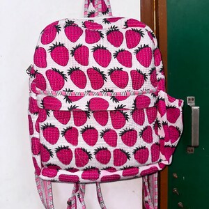 Puede incluir: Una mochila acolchada rosa y blanca con estampado de fresas. La mochila tiene correas ajustables y un peque&ntilde;o bolsillo en la parte delantera.