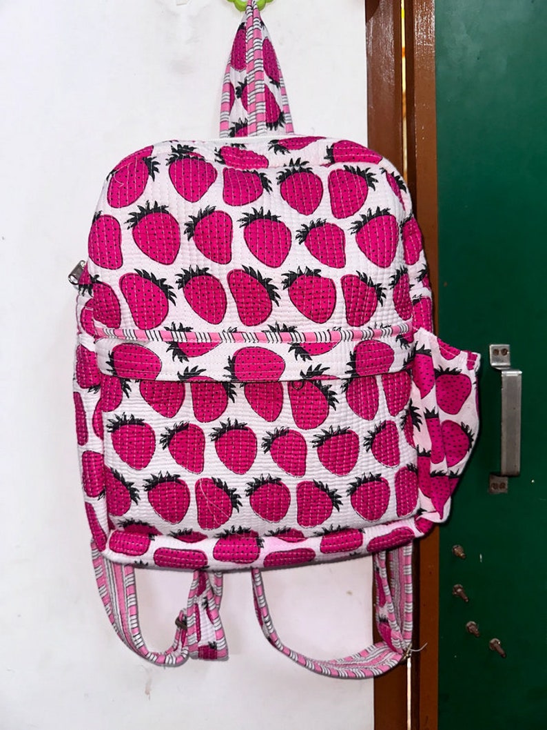 Puede incluir: Una mochila acolchada rosa y blanca con estampado de fresas. La mochila tiene correas ajustables y un peque&ntilde;o bolsillo en la parte delantera.