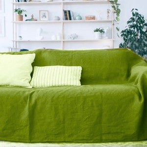 Op de afbeelding: Een groene linnen sofa deken met twee lichtgroene kussens. De deken is over een sofa met een witte achtergrond gelegd.