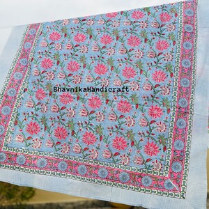 May include: A blue and pink floral patterned cotton fabric with a white border. The fabric has a repeating pattern of pink flowers and green leaves on a light blue background. The border is a white stripe with a pink and blue floral design.