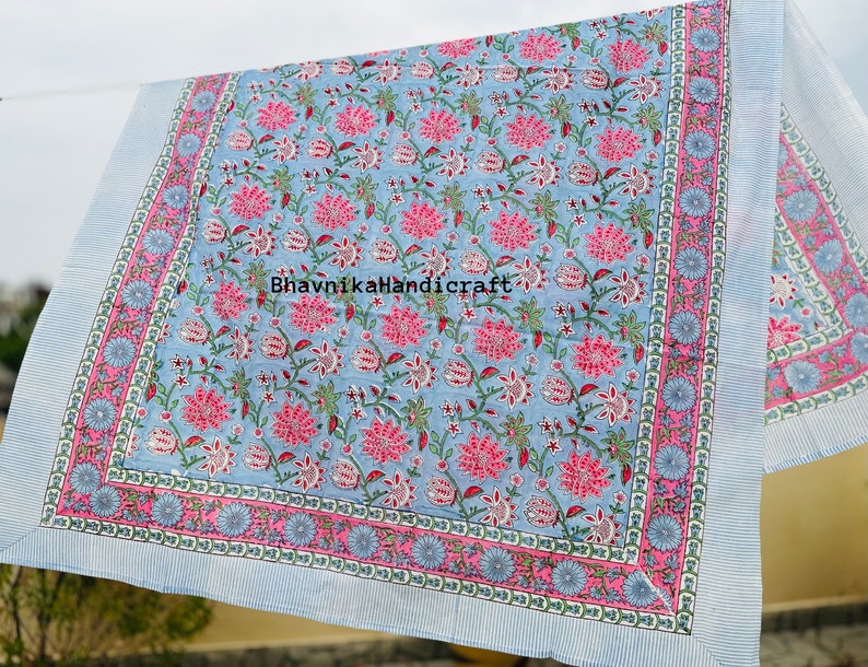 May include: A blue and pink floral patterned cotton fabric with a white border. The fabric has a repeating pattern of pink flowers and green leaves on a light blue background. The border is a white stripe with a pink and blue floral design.
