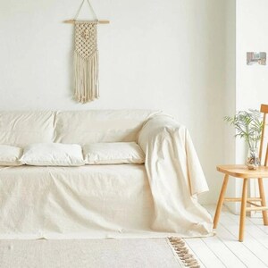 Peut inclure: Un canap&eacute; blanc avec deux coussins blancs est recouvert d'une couverture blanche. Une chaise en bois avec une plante dans un vase en verre se trouve &agrave; l'arri&egrave;re-plan. Une suspension murale en macram&eacute; est accroch&eacute;e au mur derri&egrave;re le canap&eacute;.