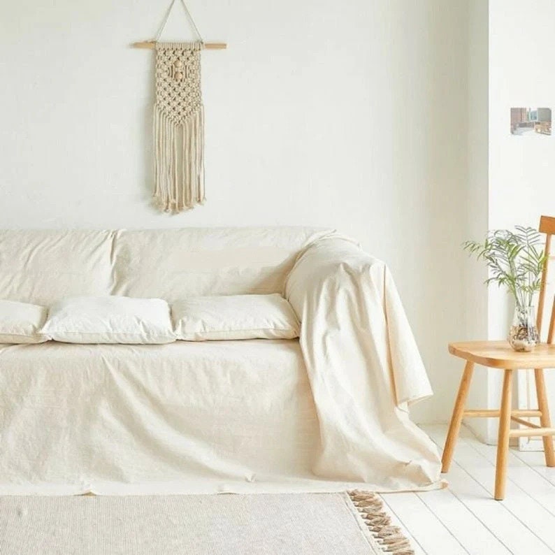 Peut inclure: Un canap&eacute; blanc avec deux coussins blancs est recouvert d'une couverture blanche. Une chaise en bois avec une plante dans un vase en verre se trouve &agrave; l'arri&egrave;re-plan. Une suspension murale en macram&eacute; est accroch&eacute;e au mur derri&egrave;re le canap&eacute;.