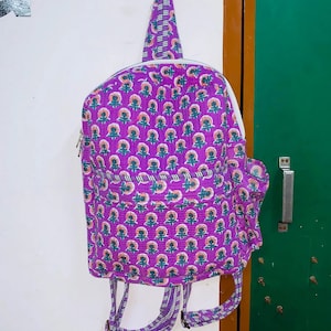 Puede incluir: Mochila morada con estampado floral y correas blancas. La mochila est&aacute; colgada de un gancho.