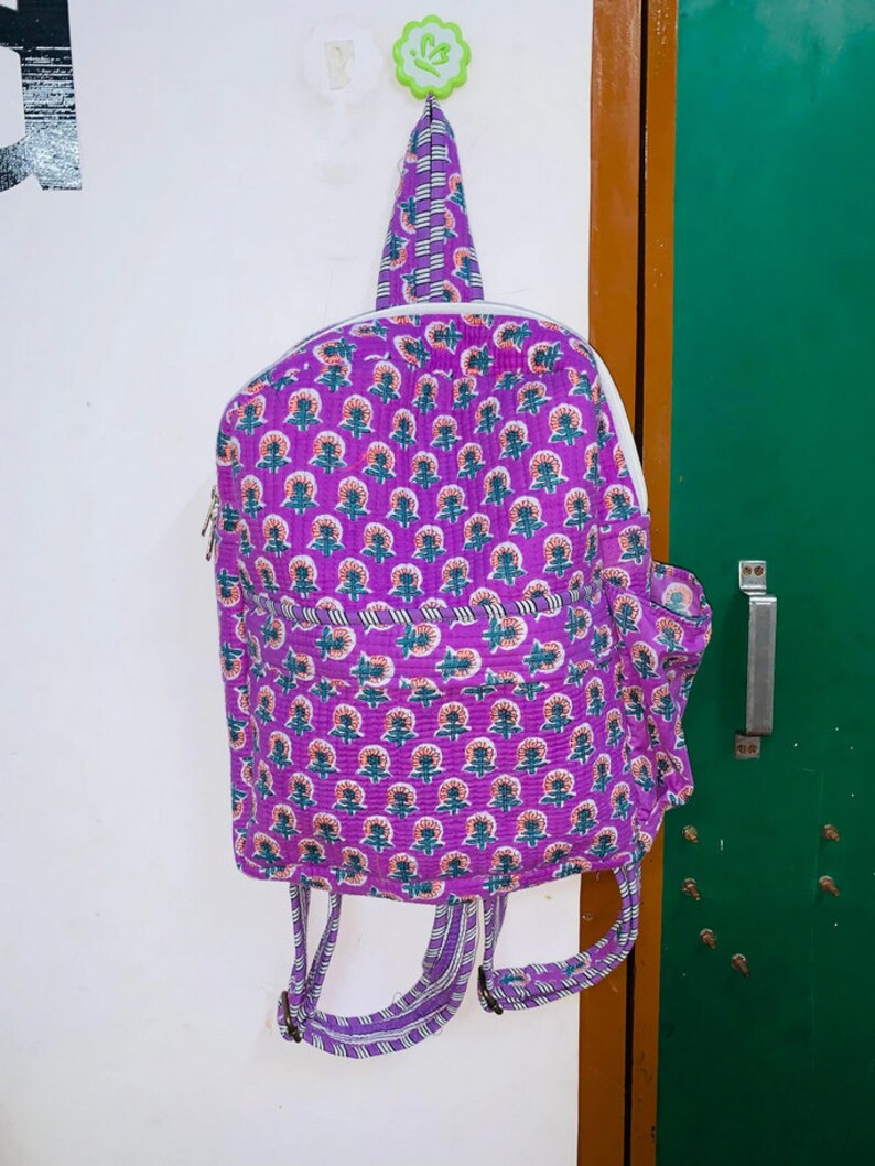 Puede incluir: Mochila morada con estampado floral y correas blancas. La mochila est&aacute; colgada de un gancho.