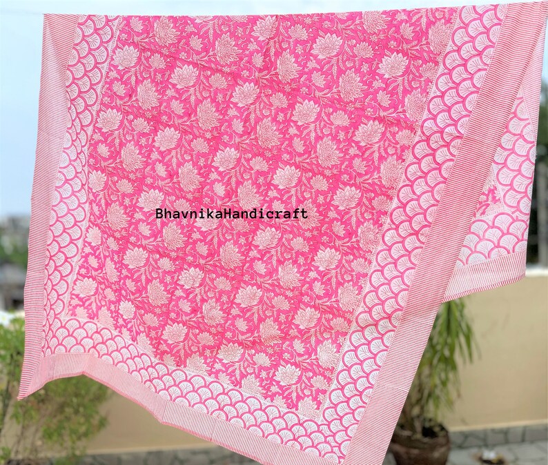 May include: Pink cotton bed sheet with a white floral pattern and a white scalloped border. The sheet is folded and hanging on a clothesline. The text "BhavnikaHandicraft" is printed in the center of the sheet.