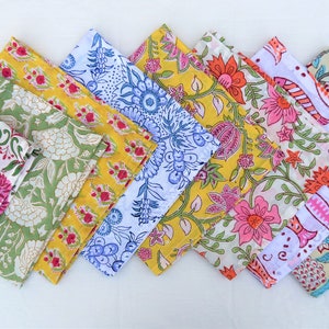 May include: A set of nine colorful cotton napkins with floral and geometric patterns. The napkins are arranged in a row, with the green napkin at the front and the pink napkin at the back.