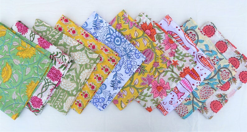May include: A set of nine colorful cotton napkins with floral and geometric patterns. The napkins are arranged in a row, with the green napkin at the front and the pink napkin at the back.