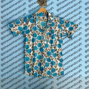 Puede incluir: Camisa de manga corta con fondo blanco y un patrón repetido de diseños florales azules. La camisa está colgada en una percha de madera. El fondo presenta el texto "@bhaveshhandicraft" repetido diagonalmente.