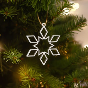 Può includere: Un ornamento natalizio bianco a forma di fiocco di neve, appeso a un filo dorato. L'ornamento presenta un design geometrico con forme a diamante e a stella. È esposto su uno sfondo di un albero di Natale verde.