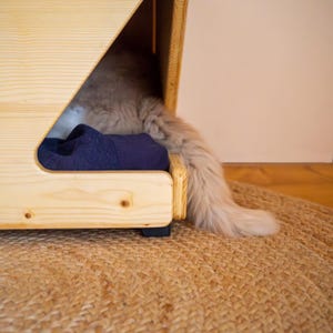 Pu&ograve; includere: Un letto per animali domestici in legno marrone chiaro con una coperta blu all'interno. Un gatto &egrave; sdraiato sul letto con la coda che pende.