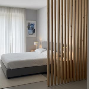 Peut inclure: Un s&eacute;parateur de pi&egrave;ce compos&eacute; de lattes de bois clair verticales dispos&eacute;es en parall&egrave;le. Le s&eacute;parateur se trouve entre un espace chambre comprenant un cadre de lit gris, une literie blanche, une table de chevet avec lampe et une &oelig;uvre murale.