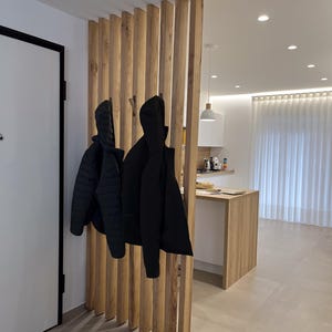 Peut inclure: Un s&eacute;parateur de pi&egrave;ce compos&eacute; de lattes de bois clair verticales, dot&eacute; de crochets int&eacute;gr&eacute;s supportant deux vestes noires. La structure sert de cloison entre une entr&eacute;e et un espace cuisine &eacute;quip&eacute; de meubles blancs et de bois clair.