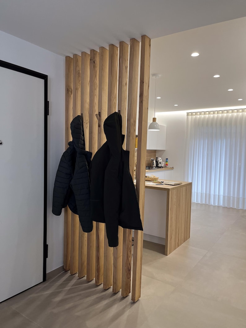 Peut inclure: Un s&eacute;parateur de pi&egrave;ce compos&eacute; de lattes de bois clair verticales, dot&eacute; de crochets int&eacute;gr&eacute;s supportant deux vestes noires. La structure sert de cloison entre une entr&eacute;e et un espace cuisine &eacute;quip&eacute; de meubles blancs et de bois clair.
